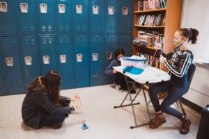 Middle School Students sitting in desk and on floor in front of blue lockers
